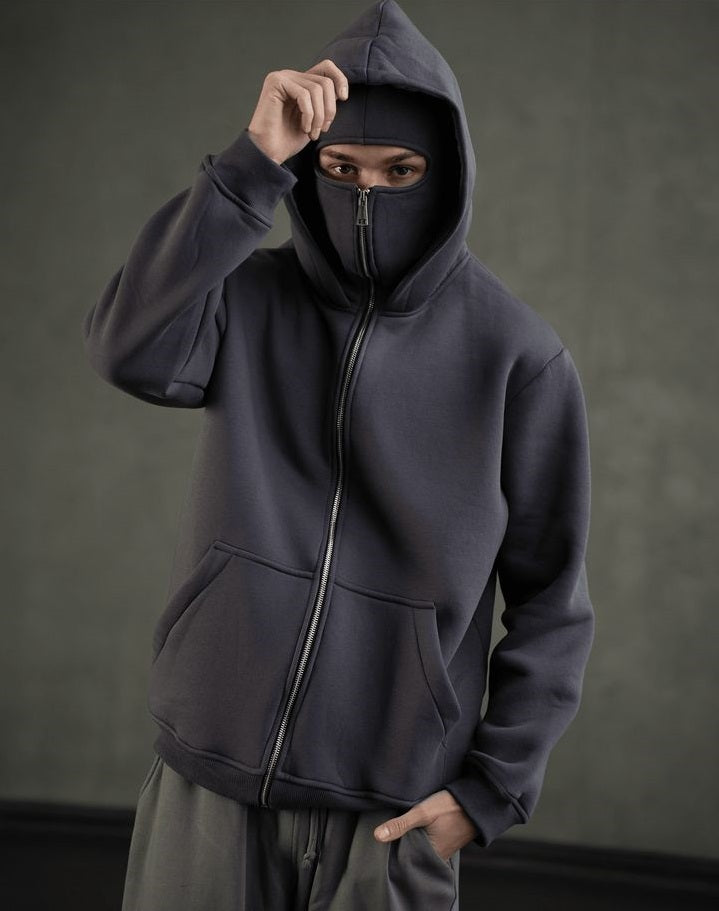Person wearing a dark gray hoodie with a face mask on a plain background